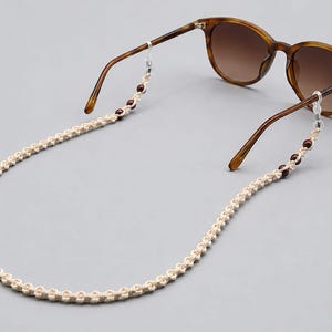 May include: Sunglasses with brown frames and gradient lenses are connected to a cream-colored macrame eyeglass chain. The chain has small, dark red beads. The glasses and chain are set against a gray background.