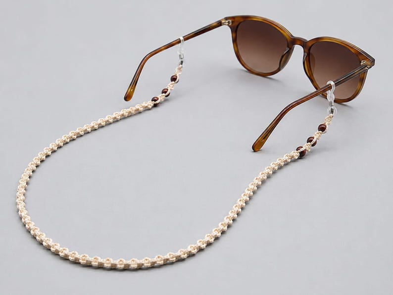 May include: Sunglasses with brown frames and gradient lenses are connected to a cream-colored macrame eyeglass chain. The chain has small, dark red beads. The glasses and chain are set against a gray background.