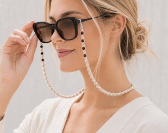 May include: Black sunglasses with a decorative chain. The chain is silver-colored with alternating dark red beads. The sunglasses have a square frame and brown-tinted lenses. The chain is attached to the arms of the sunglasses.