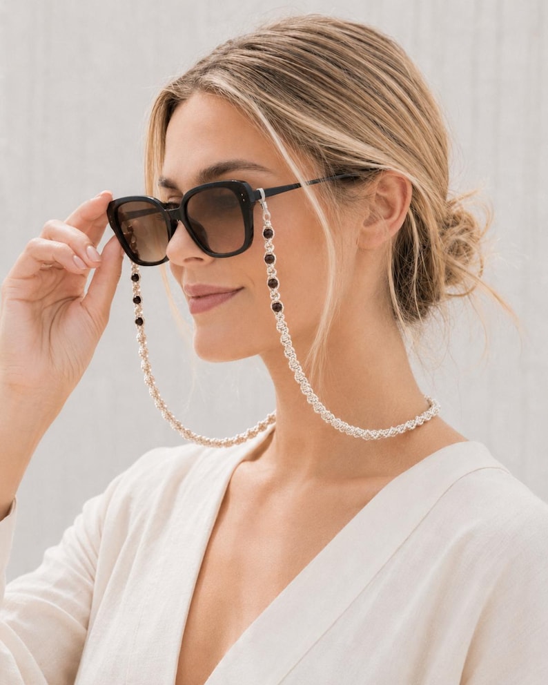 May include: Black sunglasses with a decorative chain. The chain is silver-colored with alternating dark red beads. The sunglasses have a square frame and brown-tinted lenses. The chain is attached to the arms of the sunglasses.