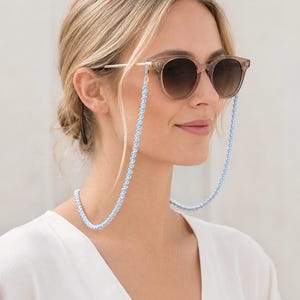 May include: Round-framed sunglasses with translucent beige frames and gradient brown lenses, featuring a light blue beaded chain attached to the temples. The chain consists of small, uniform, spherical light blue beads linked together.