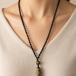 Puede incluir: Un collar de cordón negro con un colgante. El colgante es una cuenta cilíndrica amarilla y negra con un diseño intrincado. El collar se usa sobre una blusa color crema.
