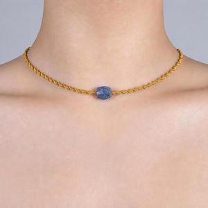Op de afbeelding: Een goudkleurige touwketting met een gefacetteerde, blauwe edelsteen hanger. De ketting is gecentreerd op de borst, waarbij het ontwerp en de kleur van de sieraden tegen de huid worden benadrukt. De ketting is een eenvoudig, elegant sieraad.