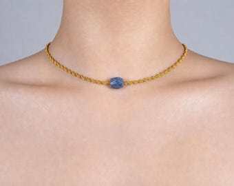 May include: A gold-colored rope chain necklace with a faceted, blue gemstone pendant. The necklace is centered on the chest, showcasing the jewelry's design and color against the skin. The necklace is a simple, elegant piece.