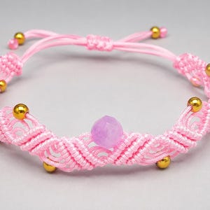 May include: A pink macrame bracelet featuring a central faceted light purple bead and multiple small metallic gold-toned spherical beads woven into the cord. The bracelet includes an adjustable sliding knot closure with two dangling cord ends.