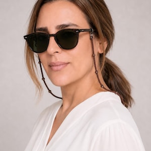 May include: Round, dark brown sunglasses with a matching brown beaded eyeglass chain. The sunglasses have a classic design and are worn by a person wearing a white blouse. The eyeglass chain is attached to the temples of the sunglasses.