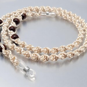 May include: A cream-colored eyeglasses chain with dark brown wooden beads. The chain is made of a twisted, knotted cord and has clear rubber loops at each end for attaching to glasses. The chain is designed to be worn around the neck.
