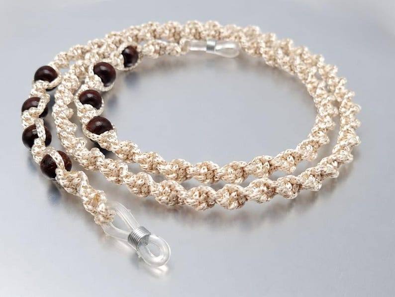May include: A cream-colored eyeglasses chain with dark brown wooden beads. The chain is made of a twisted, knotted cord and has clear rubber loops at each end for attaching to glasses. The chain is designed to be worn around the neck.