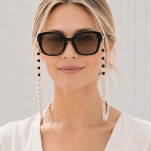 May include: Black square sunglasses with a silver chain and black bead eyeglass holder. The sunglasses have dark lenses and are worn by a person. The chain is draped around the neck.