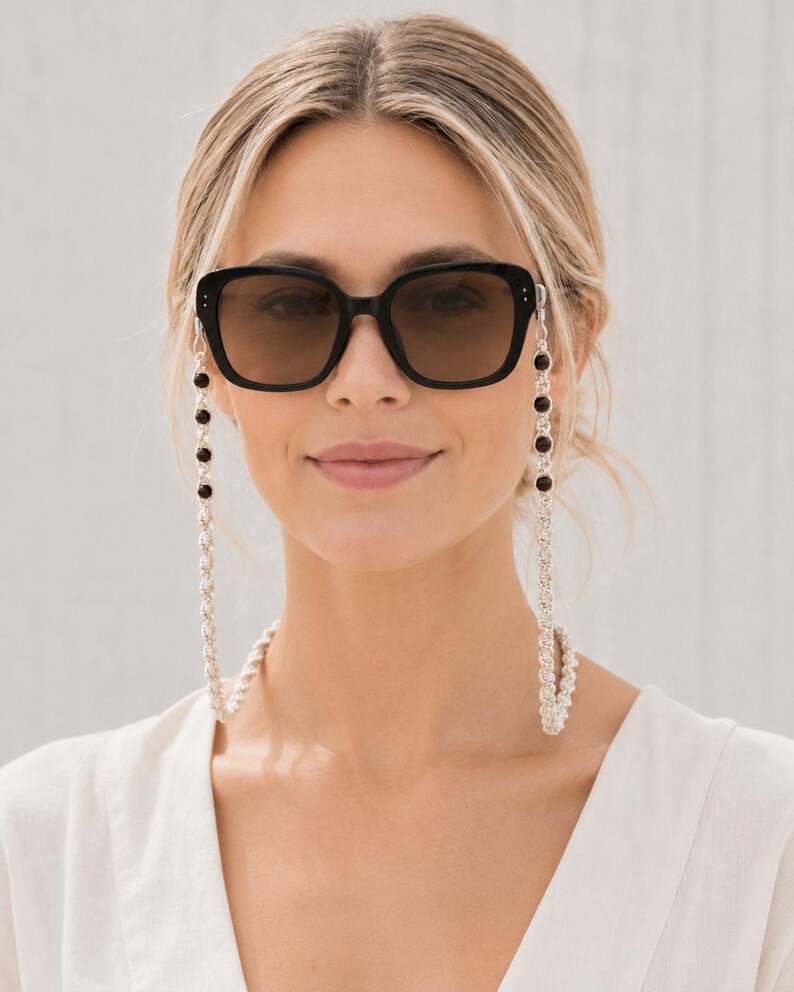 May include: Black square sunglasses with a silver chain and black bead eyeglass holder. The sunglasses have dark lenses and are worn by a person. The chain is draped around the neck.