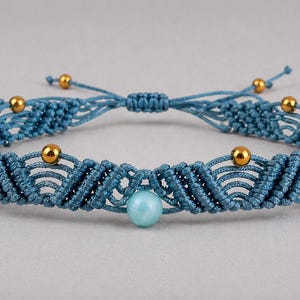 May include: A blue macrame bracelet with gold-colored beads and a central light blue bead. The adjustable bracelet features a woven design and is displayed on a gray background. The bracelet is a piece of jewelry.