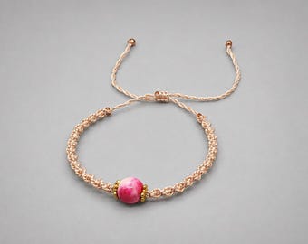 May include: A beige macrame cord bracelet featuring a single round pink and white marbled bead at the center, flanked by two small gold-toned metallic spacers. The bracelet includes an adjustable sliding knot closure with two loose cord ends.