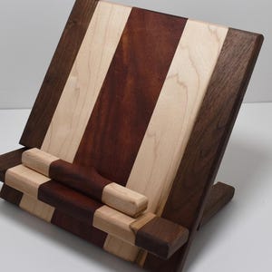 Pode incluir: Suporte de livro de receitas de madeira com um design listrado. O suporte apresenta listras alternadas de madeira marrom clara, média e escura. Possui uma borda para segurar o livro e um suporte dobrável para estabilidade.