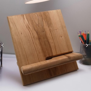 Puede incluir: Un atril de madera con acabado natural. El atril está inclinado con una repisa para sostener un libro o tableta. La veta de la madera es visible, y el atril está sobre una superficie blanca. Un portalápices con lápices de colores está en el fondo.
