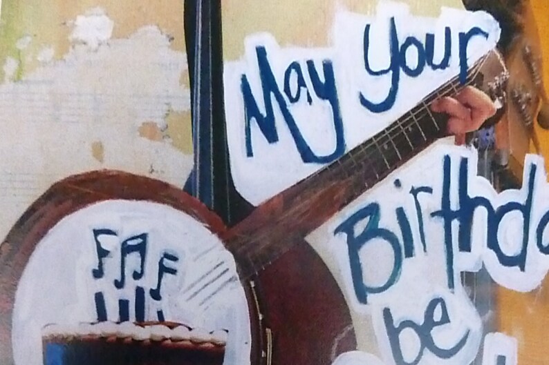 Pu&ograve; includere: Un biglietto di auguri di compleanno con sfondo bianco e lettere blu che dicono "May your birthday be". Si vede una chitarra e un banjo sullo sfondo.
