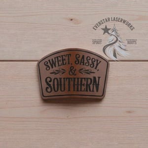 May include: A brown wooden sign with the words "SWEET, SASSY, & SOUTHERN" in black lettering. The sign has a decorative border and is set against a light wood background. The sign also has the text "EVERSTAR LASERWORKS" and a tree graphic.