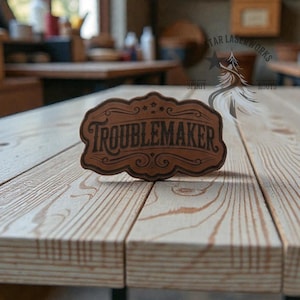 May include: A brown wooden sign with the word "TROUBLEMAKER" in a decorative font. The sign has a curved top and bottom with decorative swirls and stars. The sign is on a light-colored wooden surface.