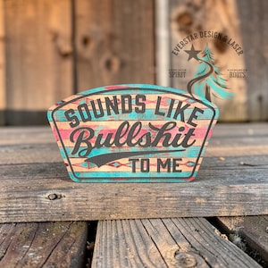 May include: Wooden sign with the phrase "SOUNDS LIKE BULLSHIT TO ME" in black lettering. The sign has a turquoise and pink patterned design with a wood grain background. The sign is in a shield shape.