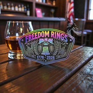 May include: A holographic sticker with the text "FREEDOM RINGS" and the years "1776-2026". The sticker features an illustration of the Liberty Bell and is placed on a wooden table next to a glass of amber liquid.