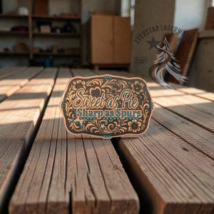 May include: A wooden belt buckle with the words "Sweet as Pie Sharp as Spurs" in a decorative font. The buckle has a floral and scroll design with turquoise accents. The background is a wooden surface.