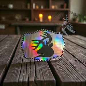 May include: A rectangular, iridescent sticker with a black deer silhouette. The sticker has a black and white striped border and is set against a wooden table. The sticker has the text "EVERSTAR FIREWORKS" and "SPIRIT" on it.