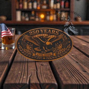 May include: Oval wooden plaque with the text "250 YEARS" and "1776-2026". The plaque features an eagle with outstretched wings, flags, and stars. The plaque is on a wooden surface with a small American flag and a glass of amber liquid in the background.