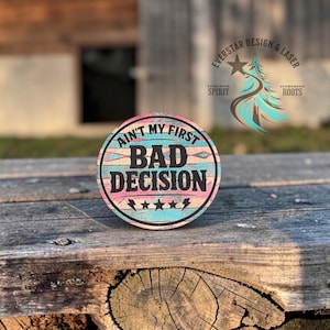 May include: A round wooden coaster with the text "Ain't My First Bad Decision" in black. The coaster has a turquoise, pink, and brown design with star and lightning bolt accents. The background is a weathered wooden surface.