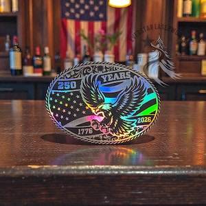 May include: An oval-shaped, laser-cut metal piece with an eagle design. The design includes the text "250 YEARS 1776-2026" and a starry section. The eagle is depicted with outstretched wings, and the piece has a holographic finish.