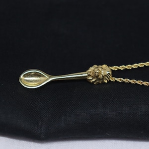 May include: A gold-colored pendant necklace featuring a miniature spoon design. The spoon has a detailed floral design at the handle's end. The necklace chain is also gold-colored, and the pendant is displayed against a dark background.
