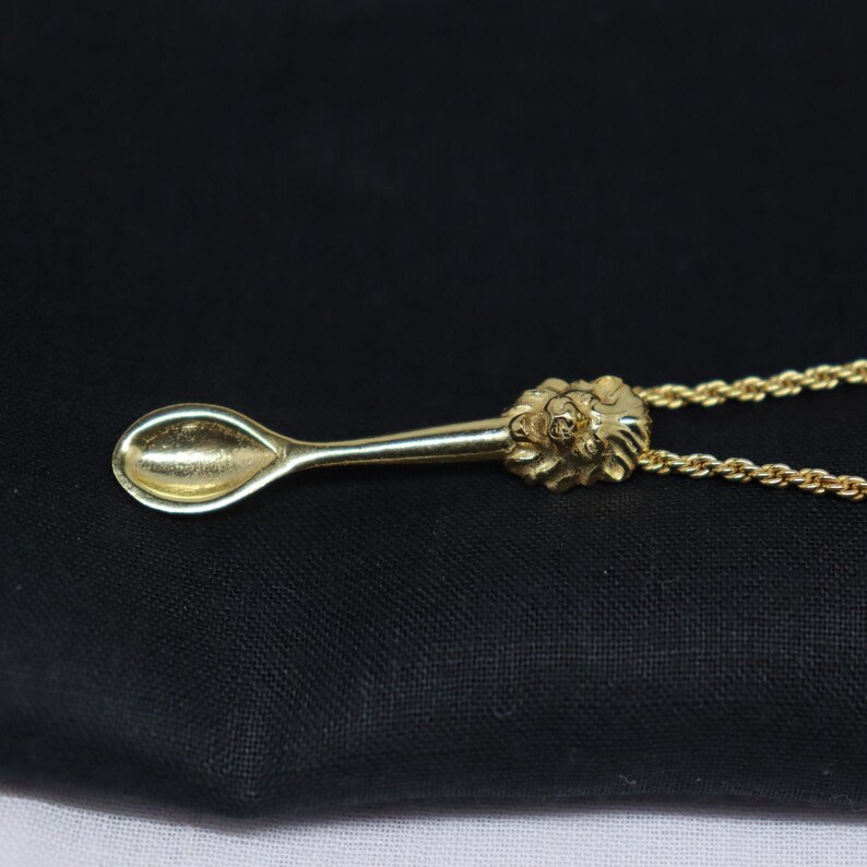 May include: A gold-colored pendant necklace featuring a miniature spoon design. The spoon has a detailed floral design at the handle's end. The necklace chain is also gold-colored, and the pendant is displayed against a dark background.