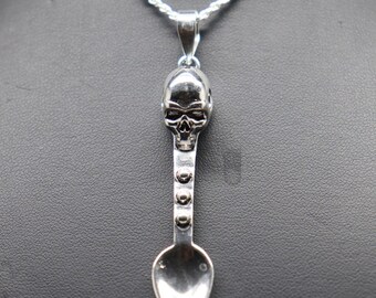 May include: A silver-toned pendant shaped like a small spoon with a skull at the top of the handle. The handle features three small circular studs arranged vertically above the spoon bowl. The pendant hangs from a silver-toned rope chain necklace.