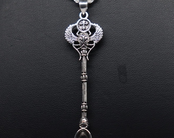 May include: A silver-toned metal spoon pendant on a matching rope chain. The handle features an Egyptian-inspired design with a winged scarab beetle motif at the top, topped by a small circular emblem containing a cross, leading to a small bowl.