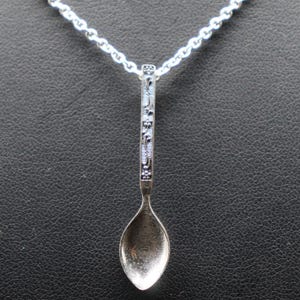 May include: A silver-toned metal spoon pendant hanging from a matching silver-toned link chain. The handle of the spoon features an intricate, embossed floral pattern with dark recessed details, while the bowl of the spoon has a smooth finish.