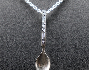 May include: A silver-toned metal spoon pendant hanging from a matching silver-toned link chain. The handle of the spoon features an intricate, embossed floral pattern with dark recessed details, while the bowl of the spoon has a smooth finish.