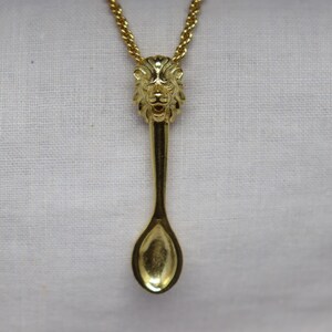 May include: A gold-colored pendant necklace featuring a spoon design. The spoon's handle is a lion's head, and the chain is a delicate gold rope. The pendant hangs against a white fabric background.