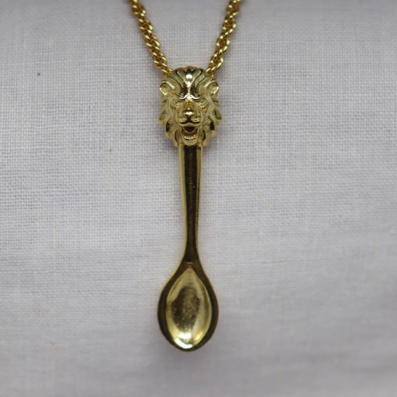 May include: A gold-colored pendant necklace featuring a spoon design. The spoon's handle is a lion's head, and the chain is a delicate gold rope. The pendant hangs against a white fabric background.