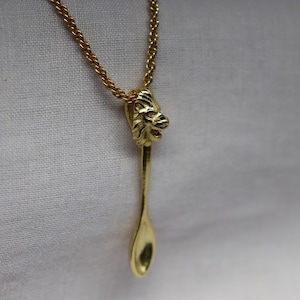 May include: Gold-toned necklace featuring a spoon-shaped pendant with a lion's head detail. The pendant hangs from a delicate chain. The spoon design adds a unique touch to this jewelry piece.