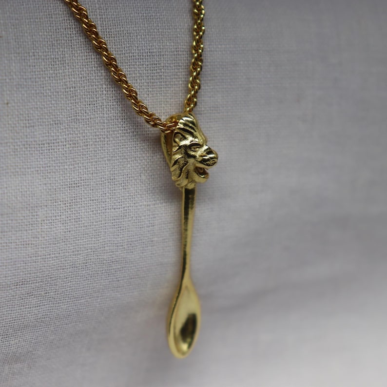 May include: Gold-toned necklace featuring a spoon-shaped pendant with a lion's head detail. The pendant hangs from a delicate chain. The spoon design adds a unique touch to this jewelry piece.