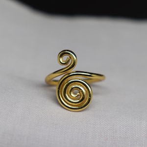 Könnte beinhalten: Ein goldfarbener Metallring mit einem markanten Spiraldesign auf der Vorderseite. Die Ringschiene besteht aus einem durchgehenden Draht, der sich zu einer kreisförmigen Spirale windet und eine glatte, polierte Oberfläche mit glänzendem Finish bildet.