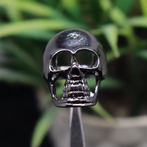 May include: A dark gray skull ring with detailed facial features. The ring has a polished finish, with eye sockets and a detailed jawline. The ring is held up against a blurred green background.