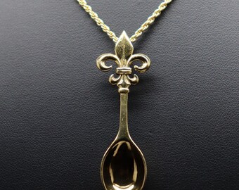 May include: A gold-toned necklace featuring a pendant shaped like a small spoon with a fleur-de-lis emblem at the top. The pendant is suspended from a thin, twisted gold-toned rope chain that extends upward from the top of the fleur-de-lis design.