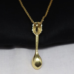 May include: A gold-colored pendant necklace featuring a spoon-shaped charm. The spoon charm has a detailed handle and a rounded bowl. The necklace chain is also gold-colored, creating a cohesive and elegant look. The pendant is displayed against a black and white background.