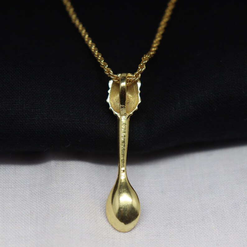 May include: A gold-colored pendant necklace featuring a spoon-shaped charm. The spoon charm has a detailed handle and a rounded bowl. The necklace chain is also gold-colored, creating a cohesive and elegant look. The pendant is displayed against a black and white background.