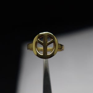 May include: A gold ring with the peace symbol design. The ring has a plain band and a circular face with the peace sign cut out. The ring is shown against a dark background.