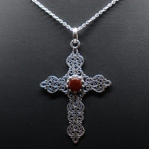 Könnte beinhalten: Halskette mit silberfarbenem Anhänger, der ein detailliertes Kreuz-Design mit Filigran aufweist. Das Kreuz hat einen ovalen, rotbraunen Stein in der Mitte. Die Kette ist filigran und der Hintergrund ist schwarz.