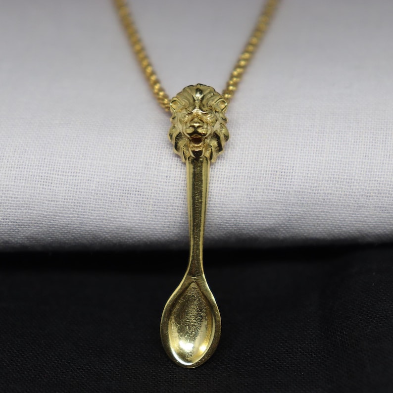 May include: A gold-colored necklace featuring a spoon pendant with a lion's head at the top. The spoon has a detailed bowl and handle, and the lion's head is intricately sculpted. The necklace chain is also gold-colored.