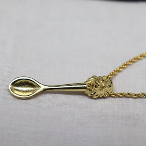 May include: A gold-colored pendant necklace featuring a spoon design. The spoon has a detailed handle with a lion's head and is suspended from a delicate chain. The spoon's bowl is oval-shaped.