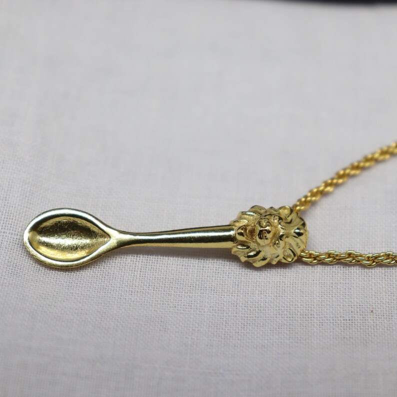 May include: A gold-colored pendant necklace featuring a spoon design. The spoon has a detailed handle with a lion's head and is suspended from a delicate chain. The spoon's bowl is oval-shaped.