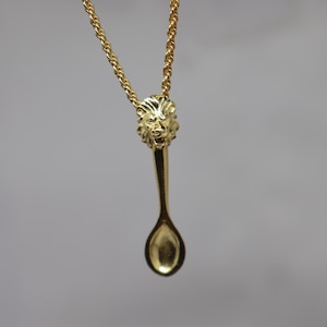 May include: A gold-colored necklace featuring a spoon-shaped pendant. The pendant has a lion's head at the top and a spoon bowl at the bottom. The chain is a twisted rope design, and the background is a solid gray.