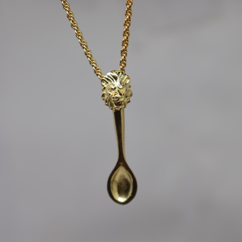 May include: A gold-colored necklace featuring a spoon-shaped pendant. The pendant has a lion's head at the top and a spoon bowl at the bottom. The chain is a twisted rope design, and the background is a solid gray.
